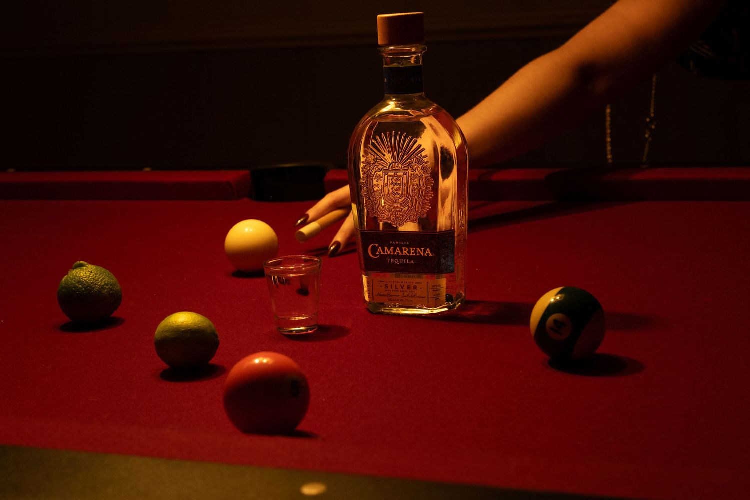 A bottle of tequila sits on a red pool table surrounded by scattered billiard balls in moody lighting.