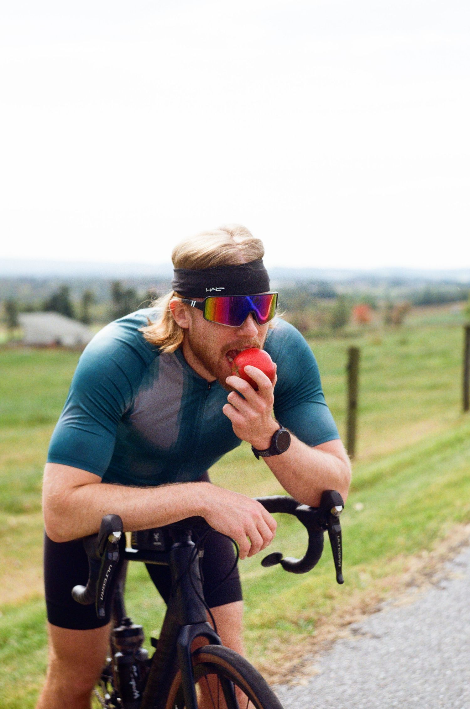 A cyclist in a blue jersey takes a break while resting on their bike handlebars on a rural gravel road.