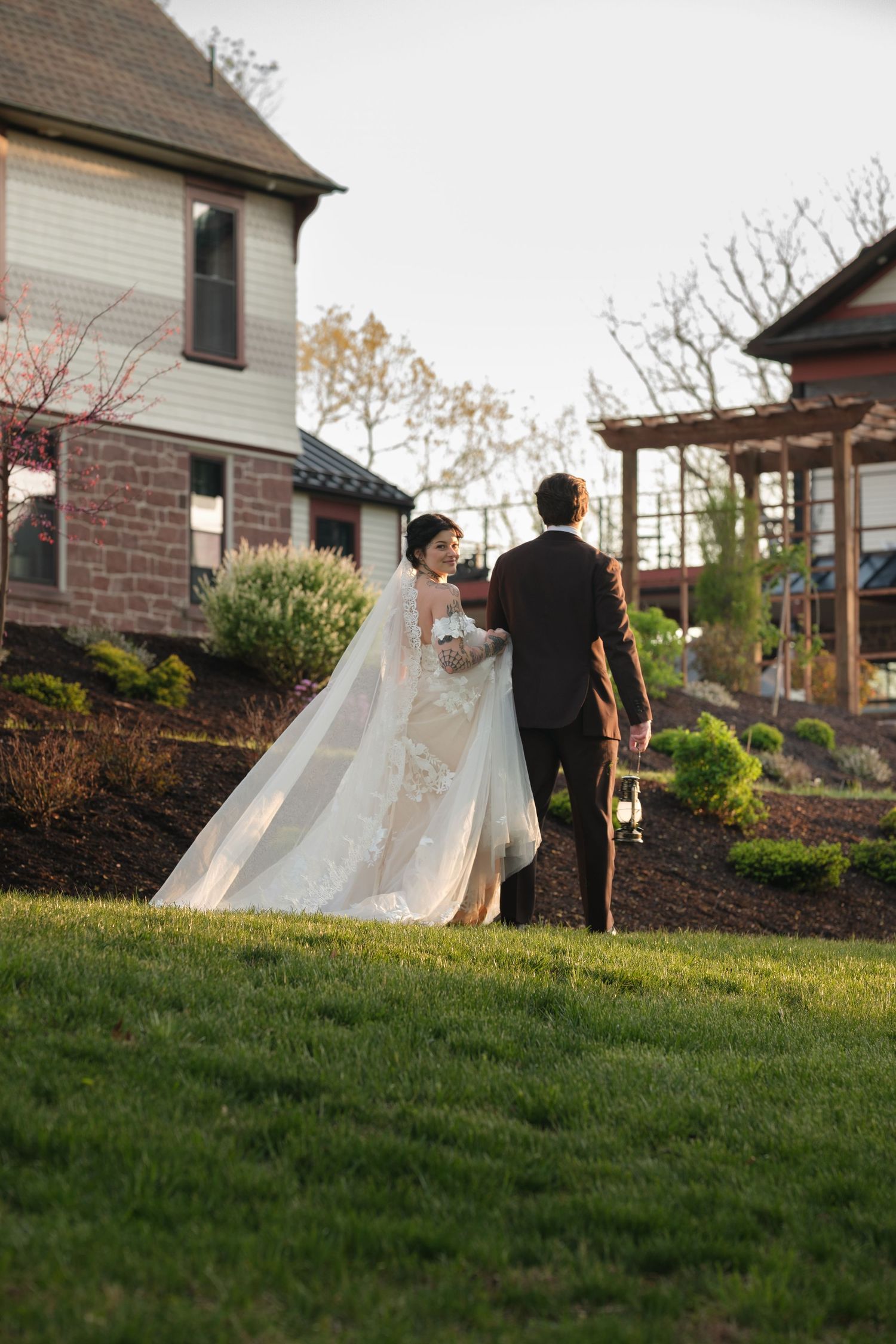 Bride in lace gown and veil walks with groom in dark suit through green garden grounds near historic stone building.