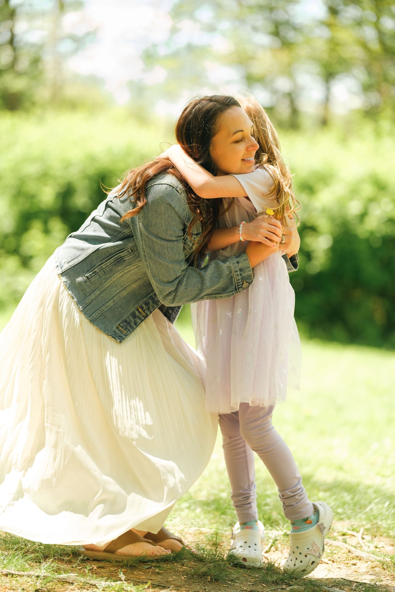 A woman in a denim jacket and white skirt hugs a young girl in a pink tutu outdoors in a sunny green field.