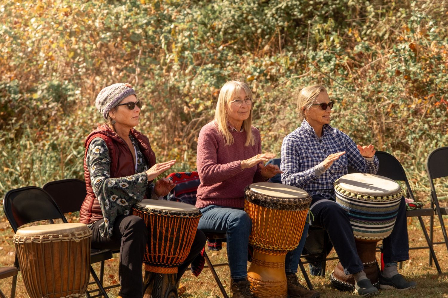 A row of senior residents sit in wooden chairs outdoors enjoying activities in an autumn garden setting.
