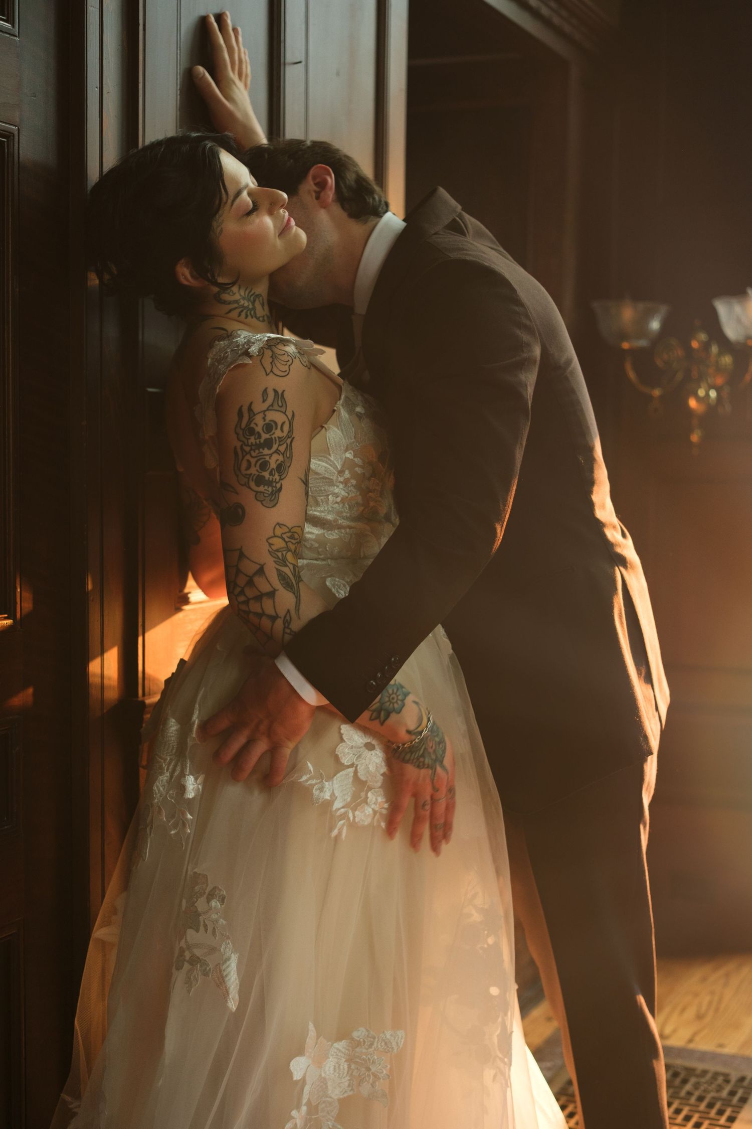 Tattooed bride in floral gown shares romantic kiss with groom in dark suit against wooden door in warm light.