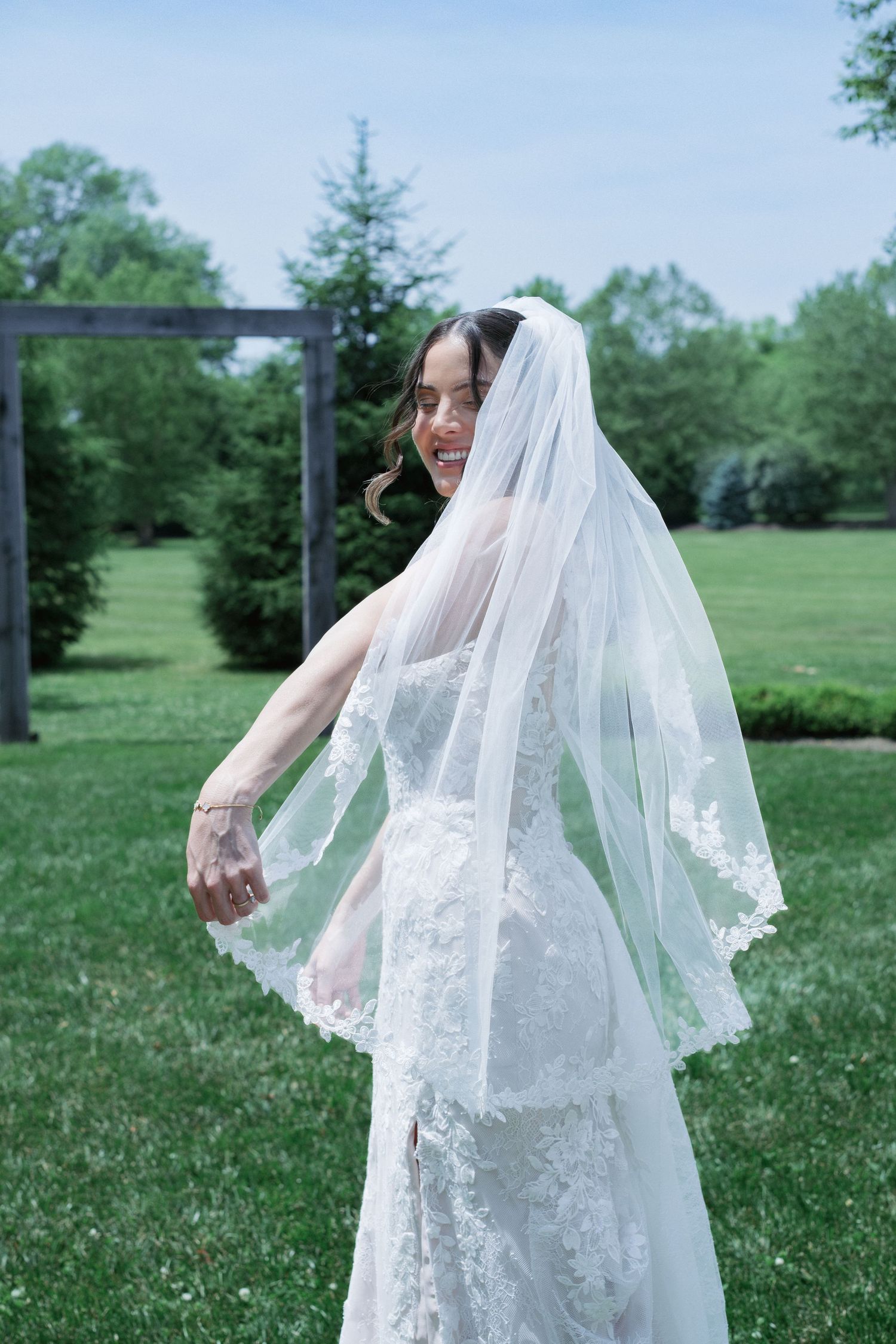 Bride in lace wedding gown and two-tier veil twirls joyfully in a lush green garden on her wedding day.