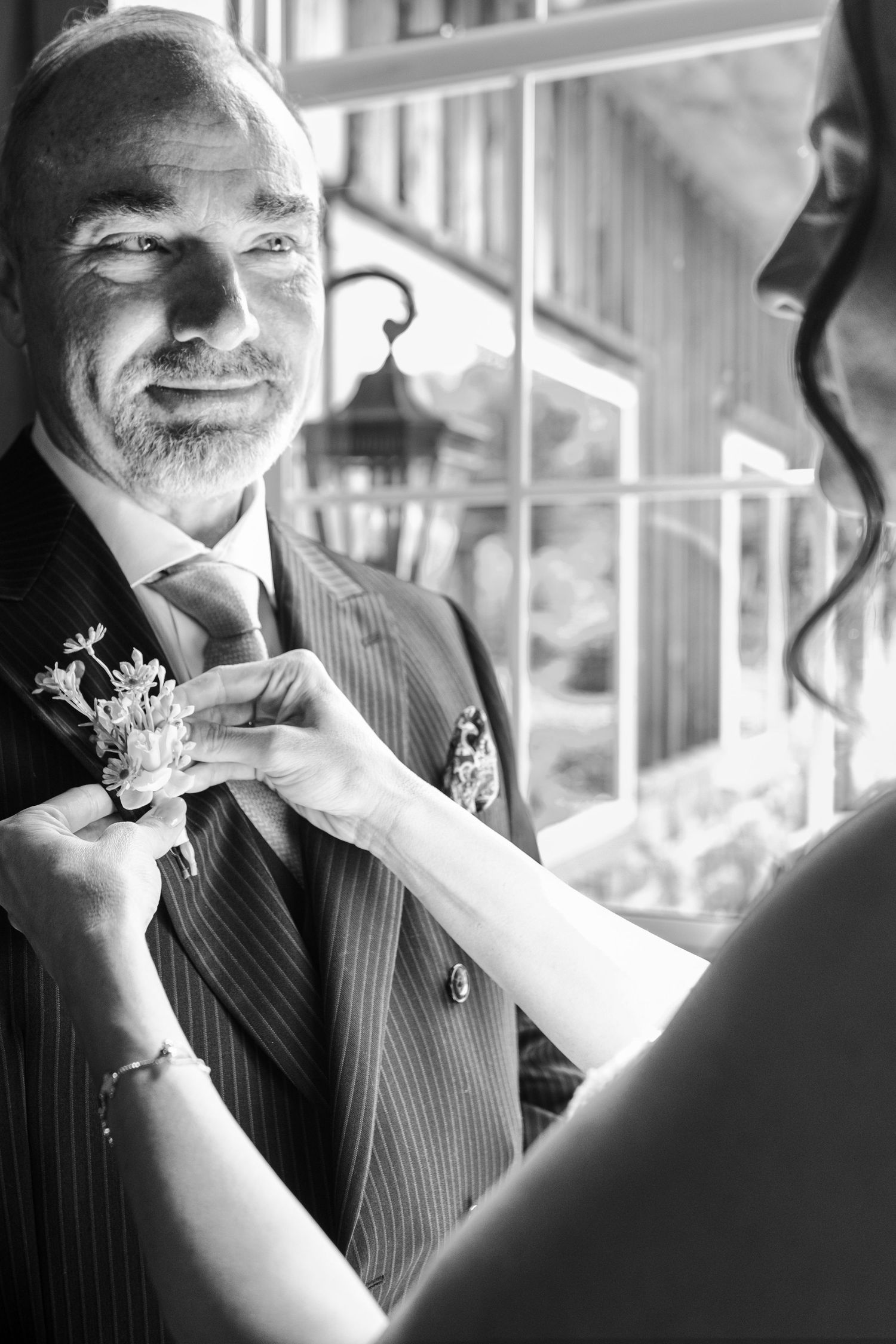 Black and white photo of smiling groom in pinstripe suit having boutonniere pinned on lapel.