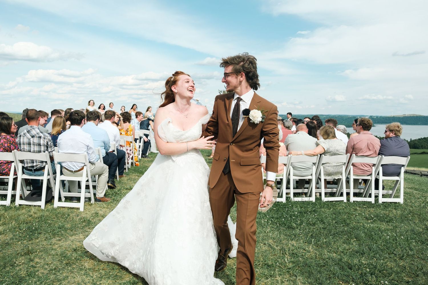 Joyful newlyweds walk down outdoor aisle after wedding ceremony, bride in white gown, groom in brown suit.