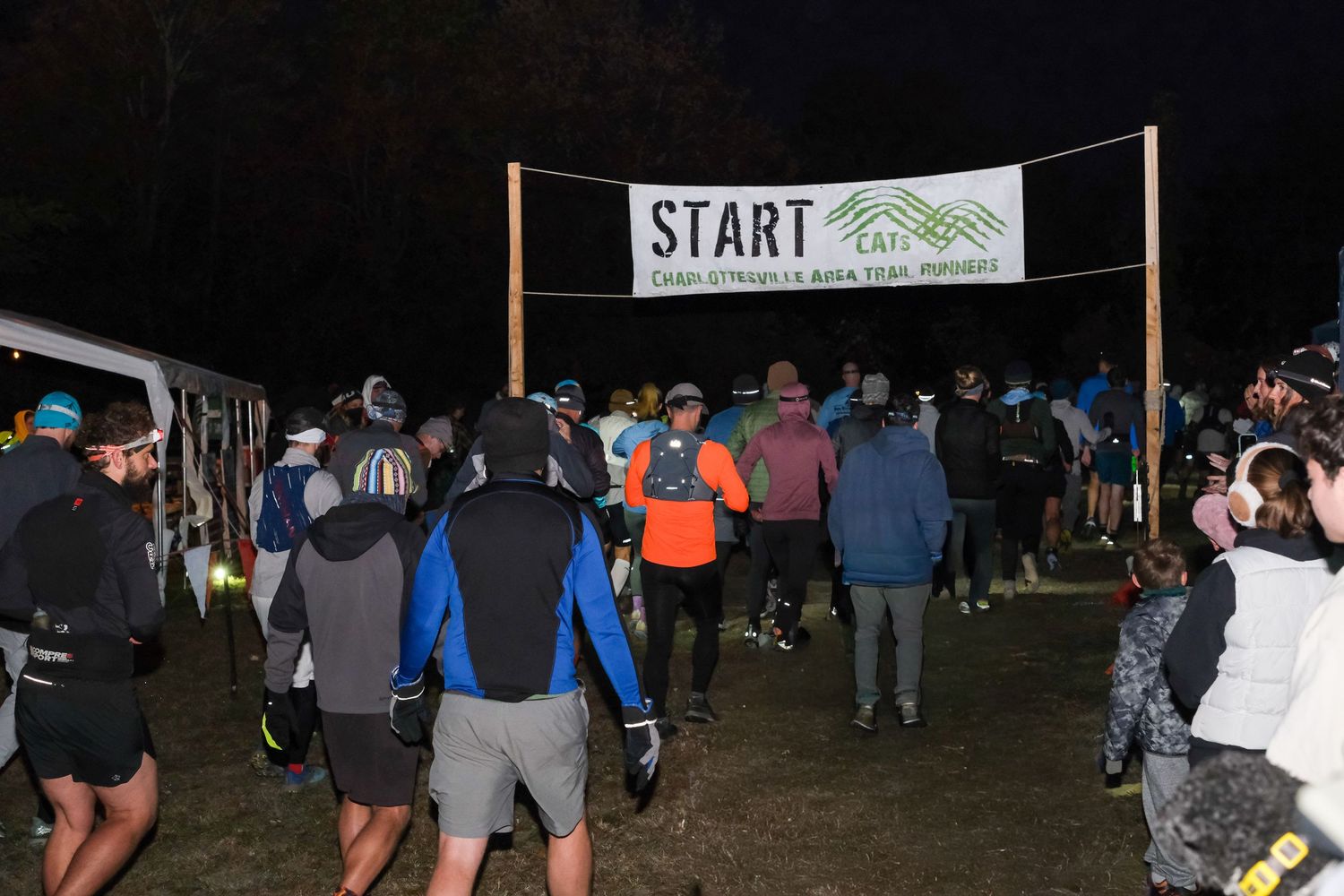 A group of runners gather at a nighttime race starting line with a banner overhead.