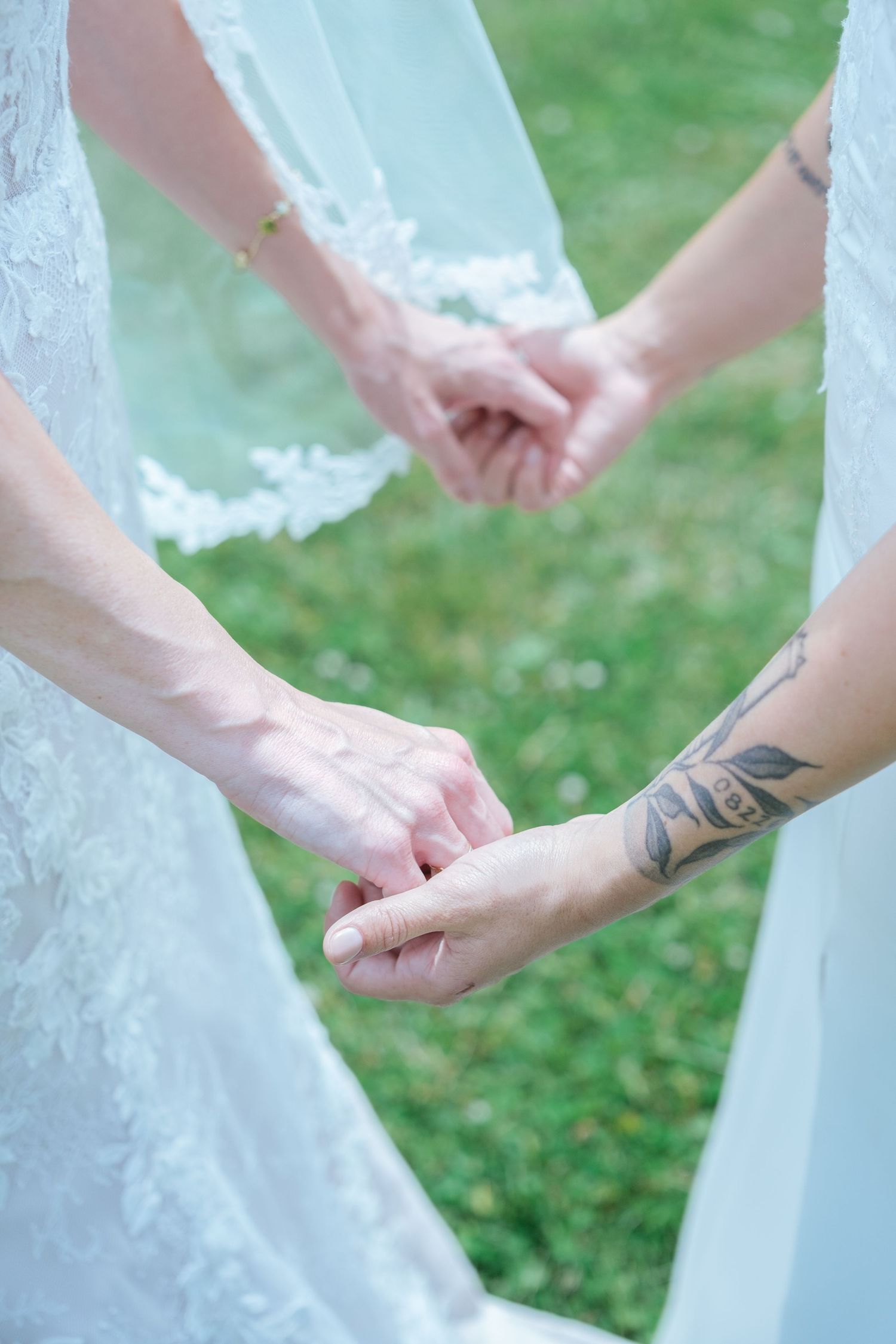 Two brides in lace wedding dresses hold hands, one with a floral tattoo on her wrist, on green grass.