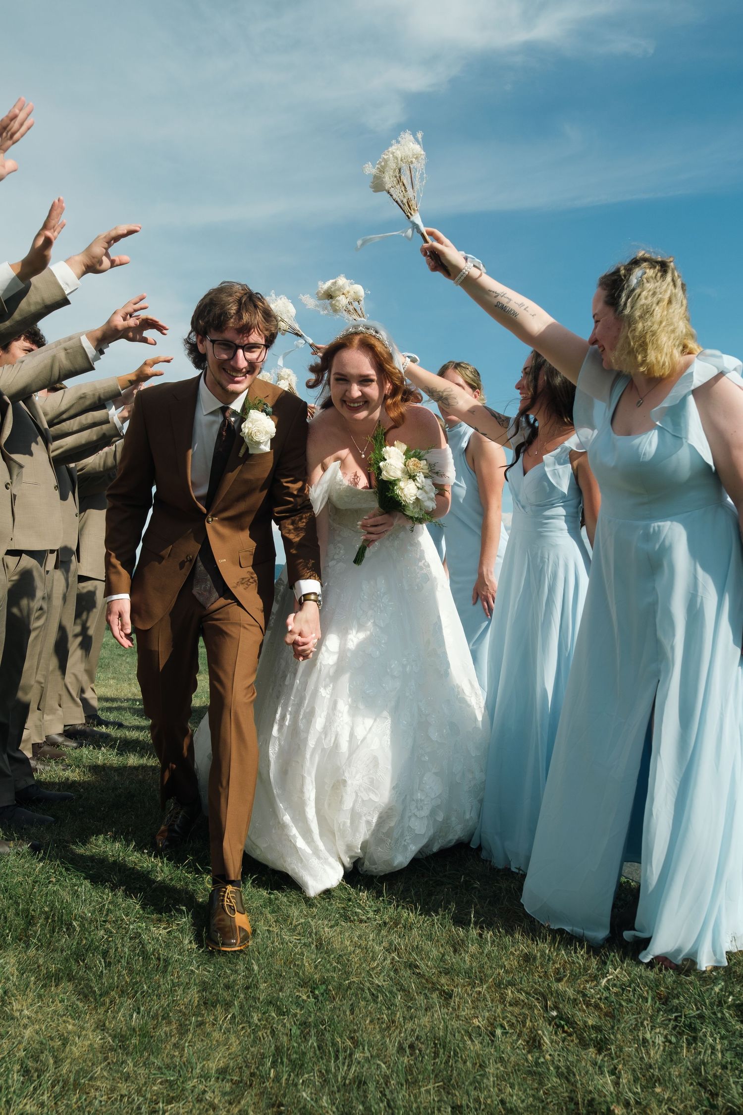 Bride and groom walking through cheering wedding party under blue sky.