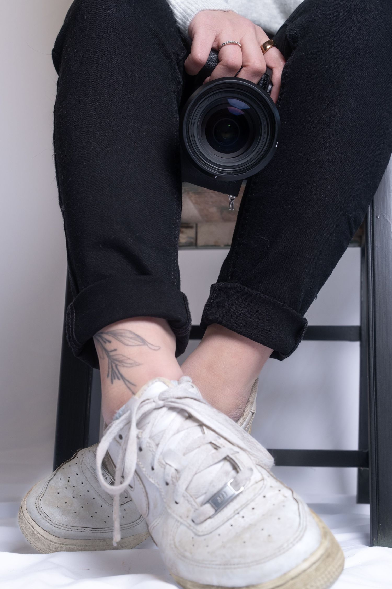 Person wearing white sneakers and rolled up black pants holding a camera lens while sitting on stairs.