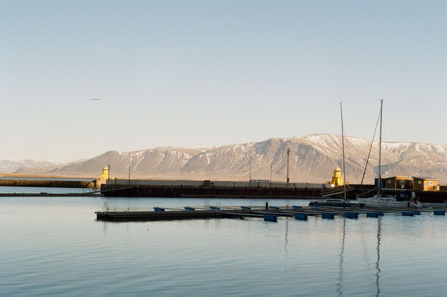 Panoramic view of a tranquil harbor with boats at sunset and snow-capped mountains in the background.