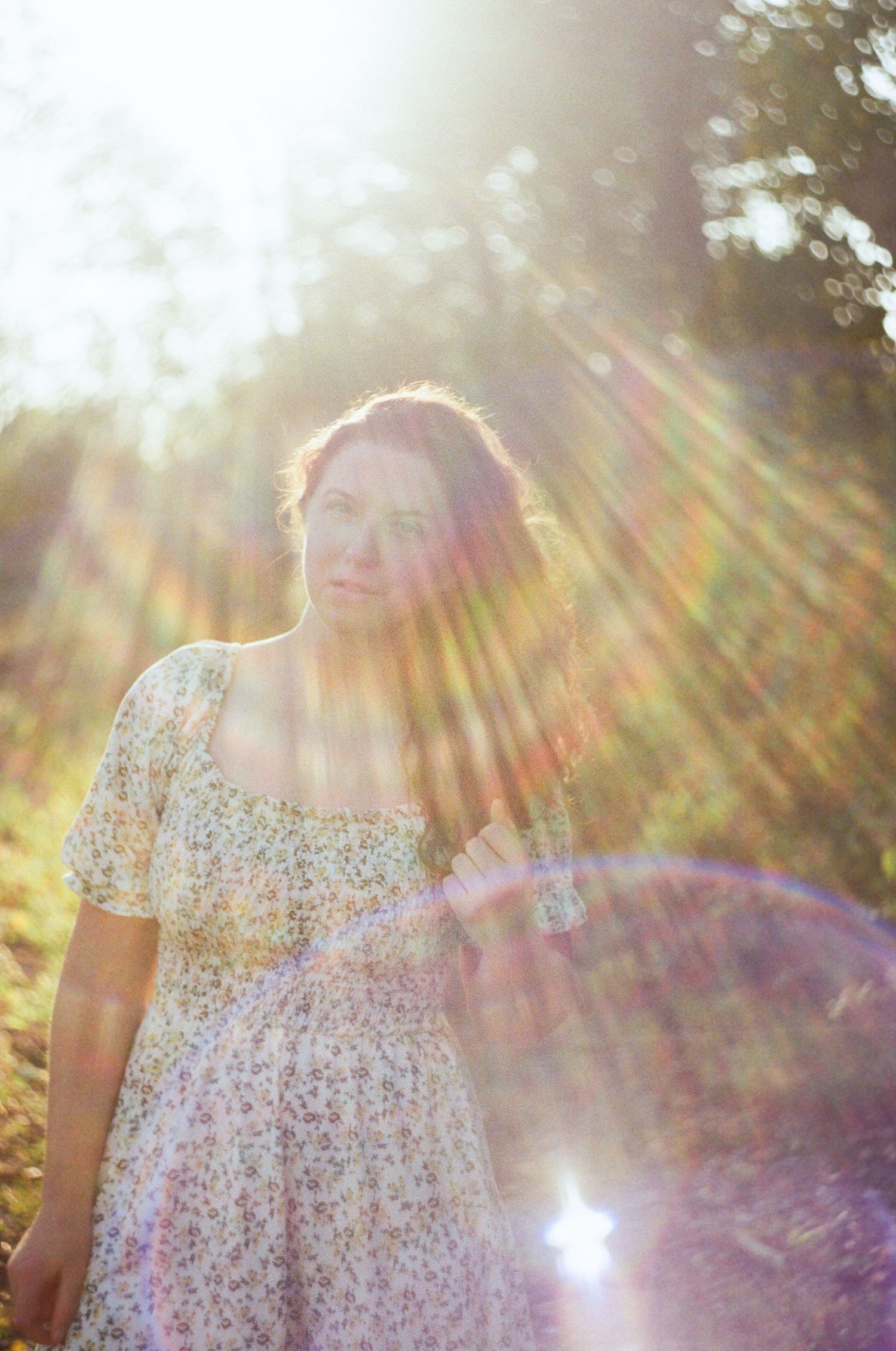 Woman in floral dress standing outdoors with sunlight rays and lens flare creating a dreamy, ethereal atmosphere.