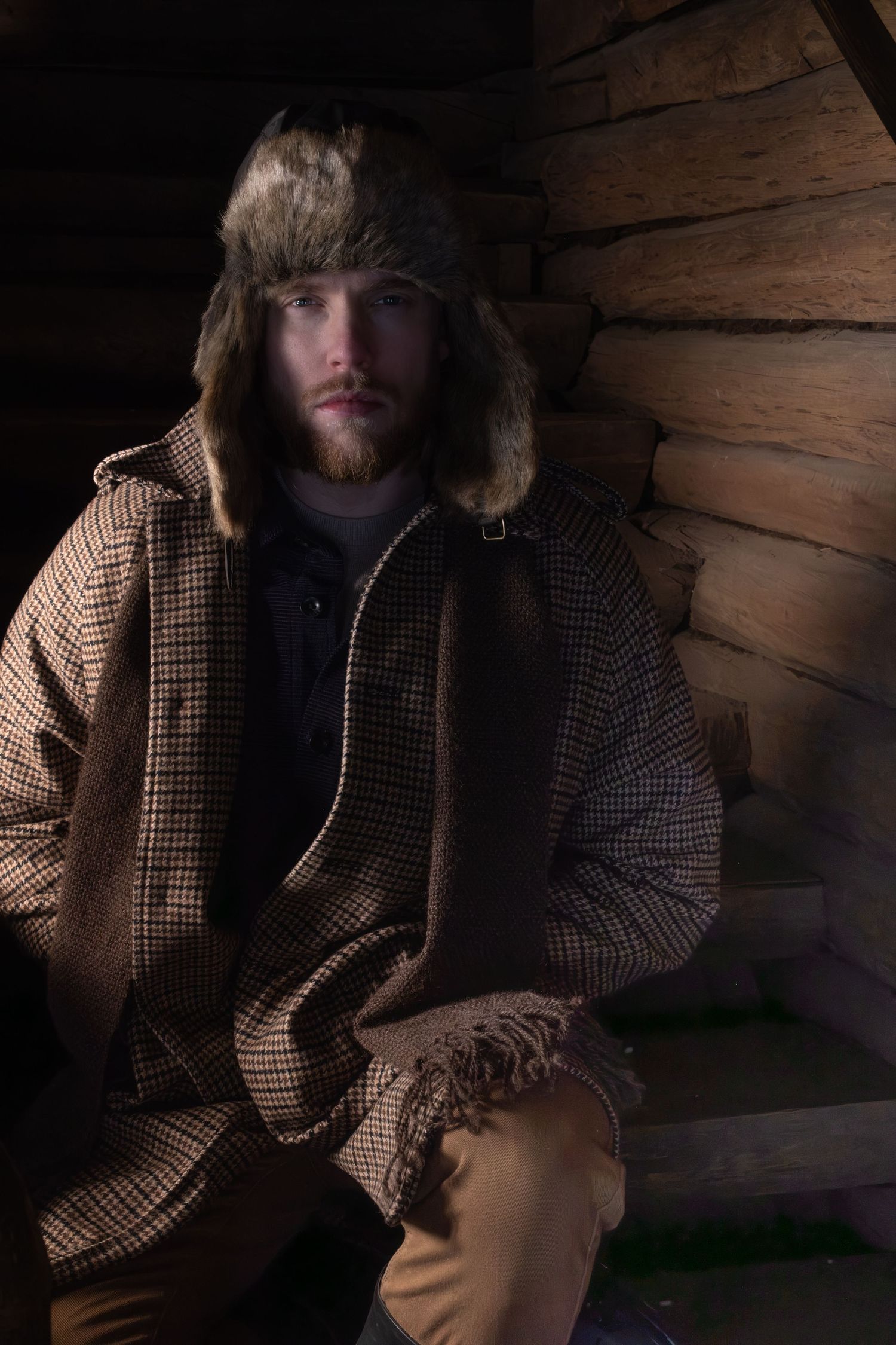 Person in plaid winter coat and fur hood sits in shadowy wooden cabin interior.