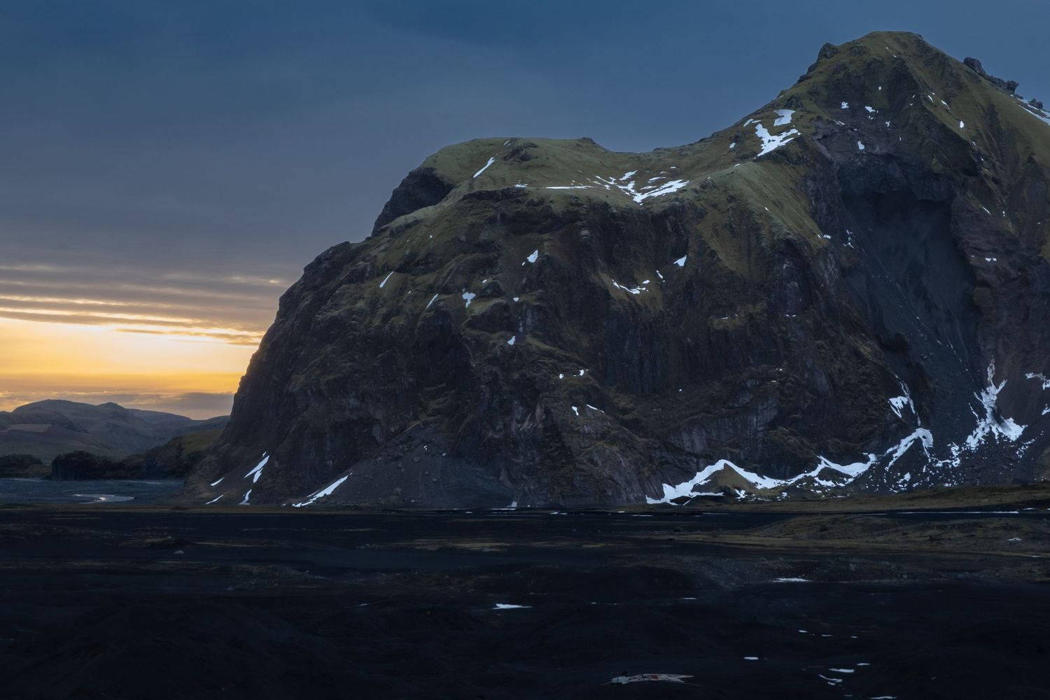 Panoramic mountain landscape with snow-capped peaks and dramatic sunset sky over rugged alpine terrain.