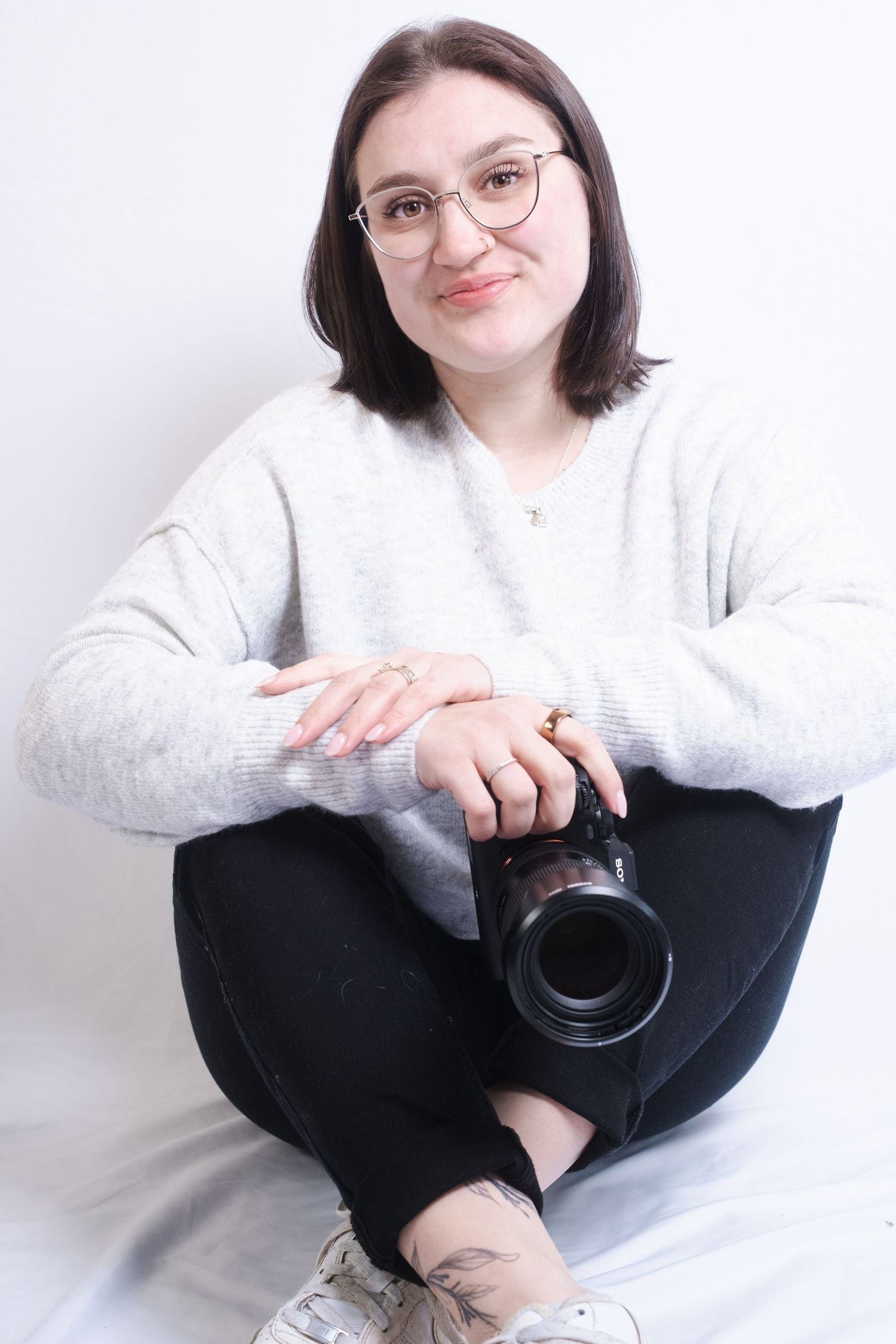 A person in a white sweater and black pants sits casually against a white wall while holding a camera lens.