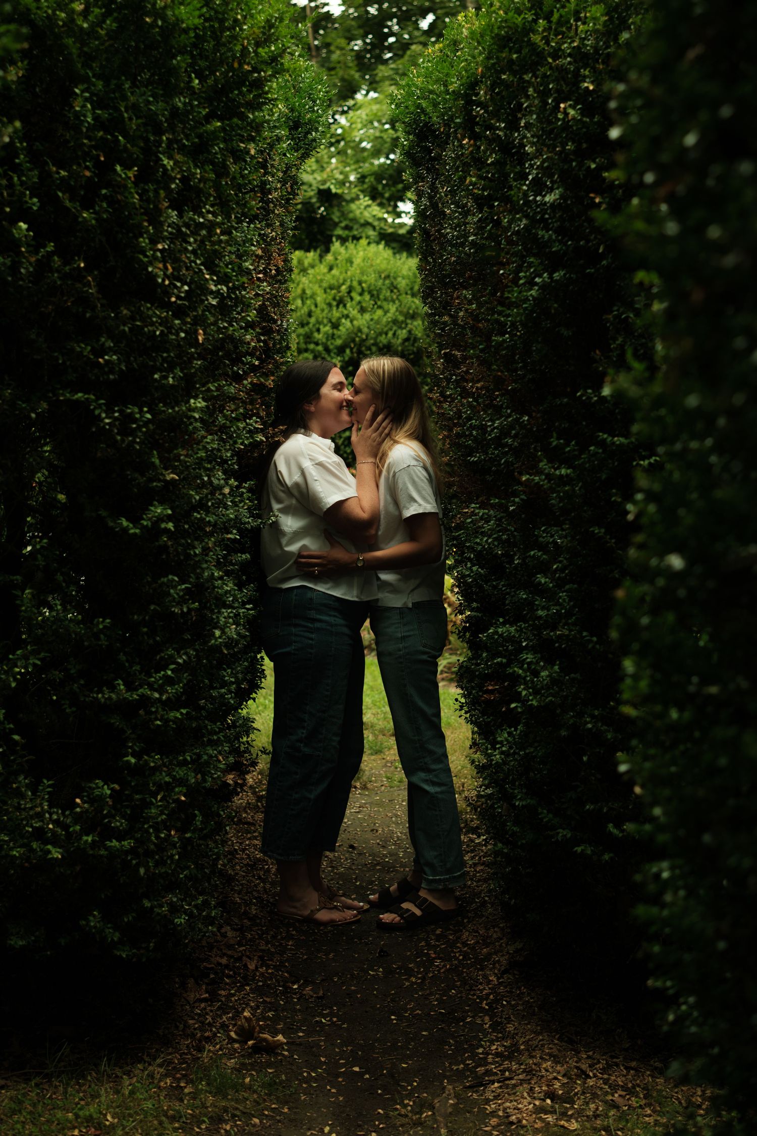 Two women sharing a romantic kiss in a lush green garden hedge maze pathway.