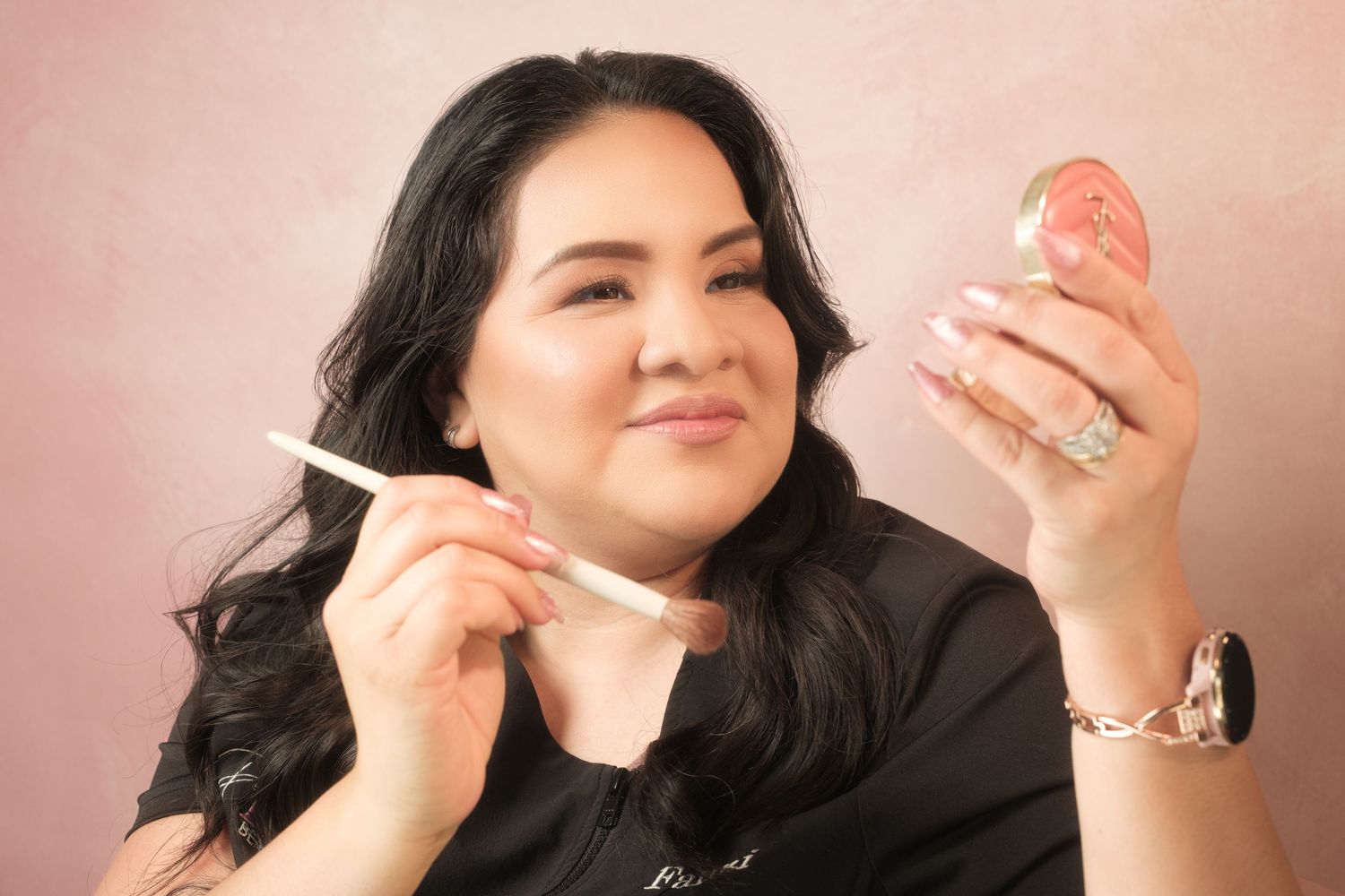 Woman applying lip gloss with a brush while holding a compact mirror, wearing a black top and jewelry.