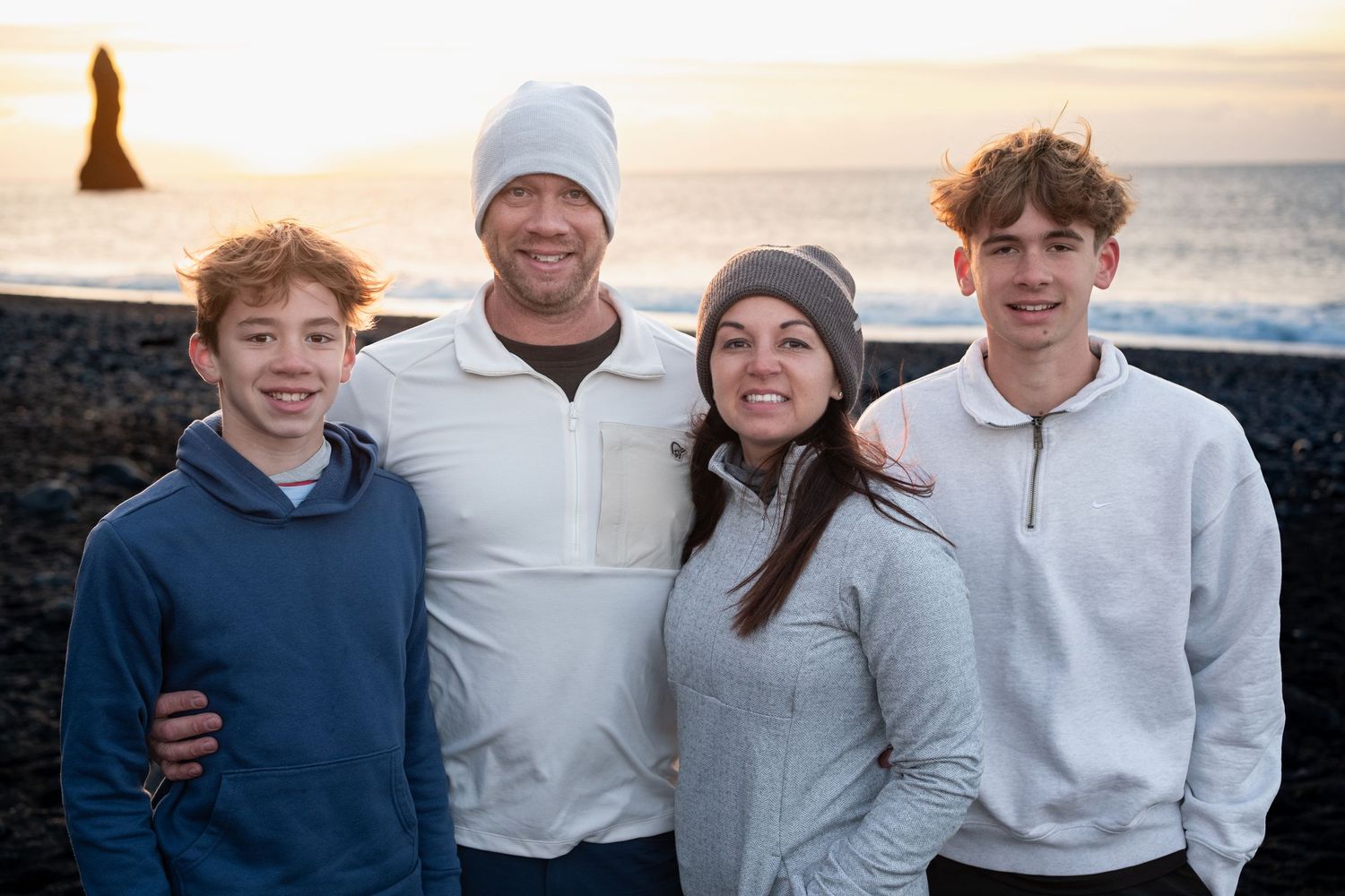 A family poses together on a beach at sunset wearing casual winter clothing in light neutral colors.