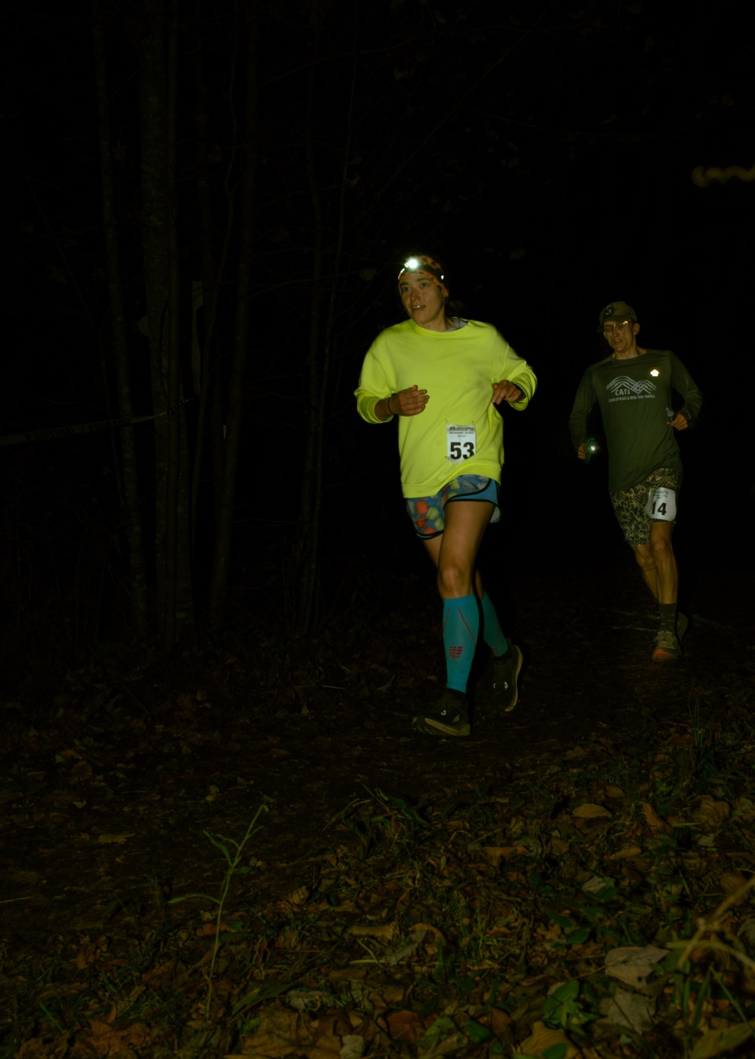 Runners wearing race bibs navigate a dark trail at night with bright neon clothing and headlamps.