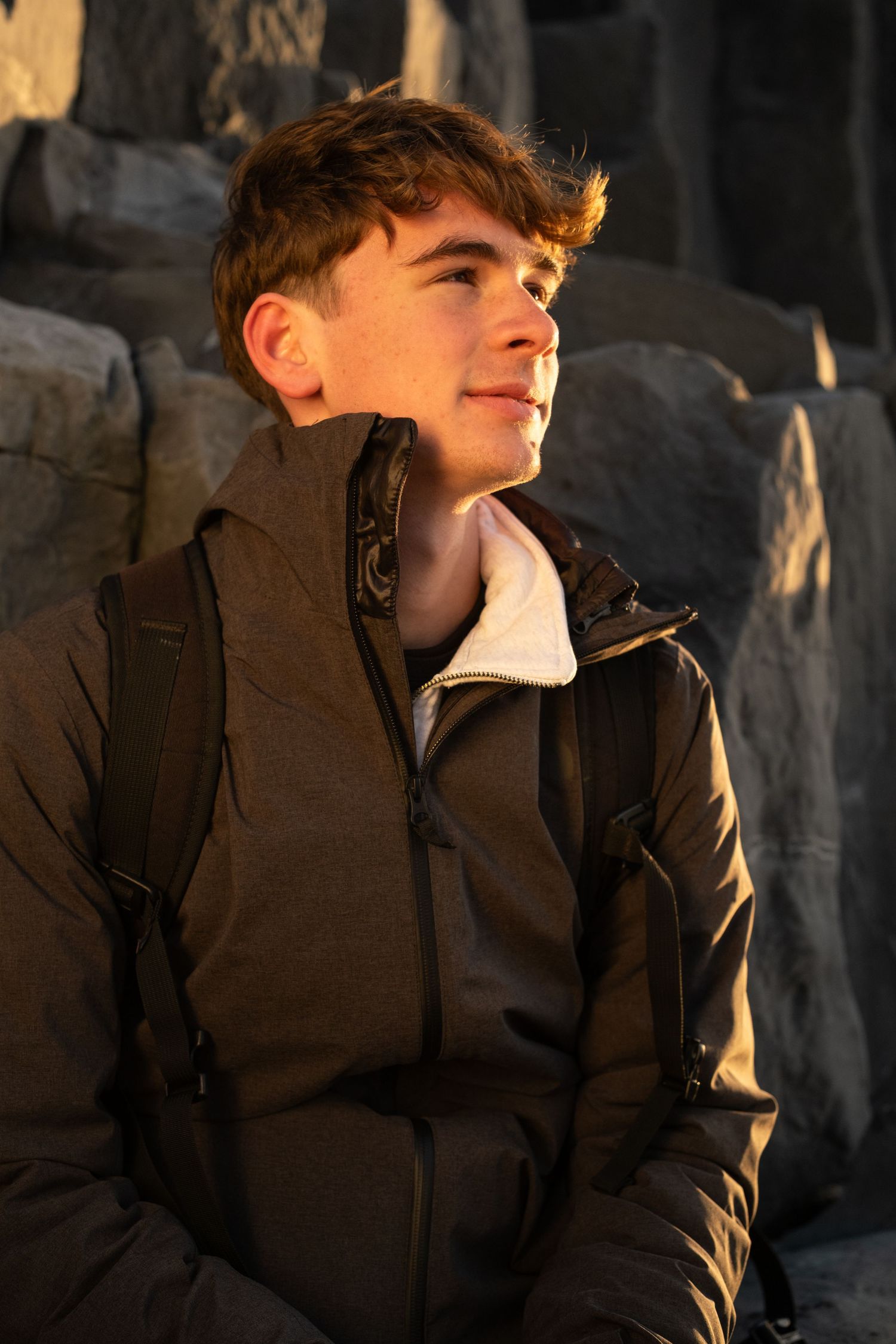 A person in a dark winter coat gazes upward while sitting against a rocky backdrop in warm lighting.