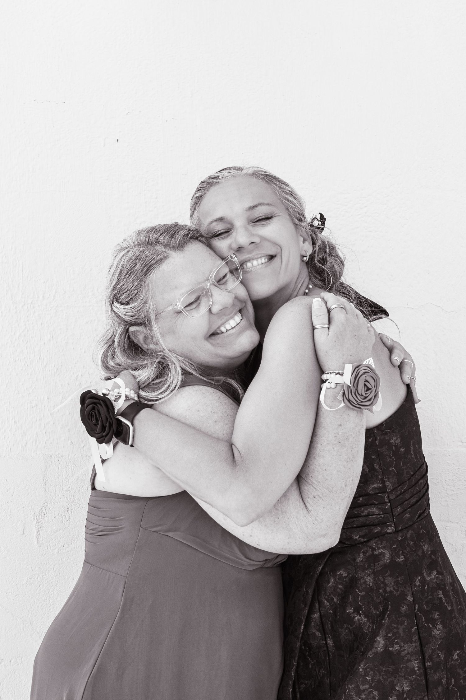 Two women in formal dresses share a warm embrace, smiling joyfully at a wedding celebration.