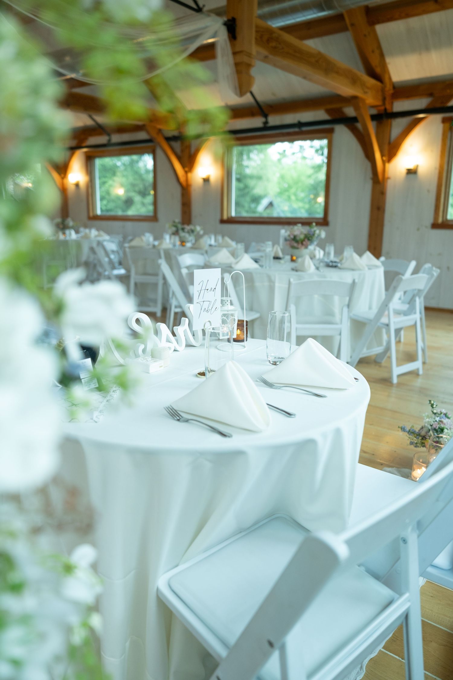 Elegant wedding reception hall with white round tables, folding chairs, floral centerpieces, and wooden beam ceiling.