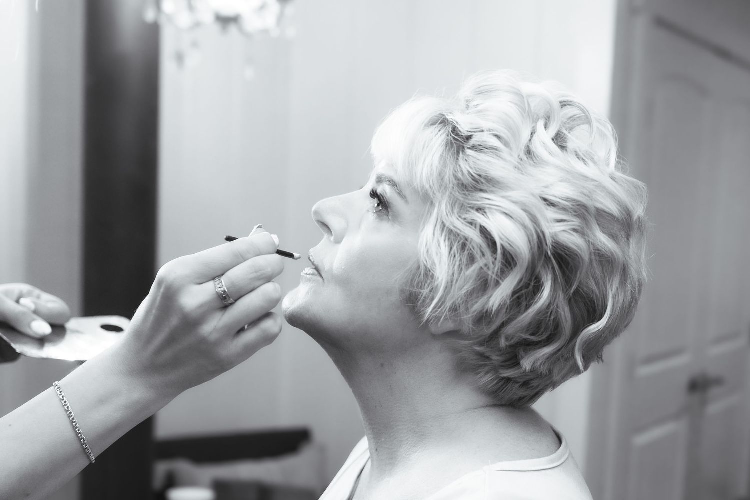 Black and white photo of a makeup artist applying lipstick to a blonde woman with short curly hair.