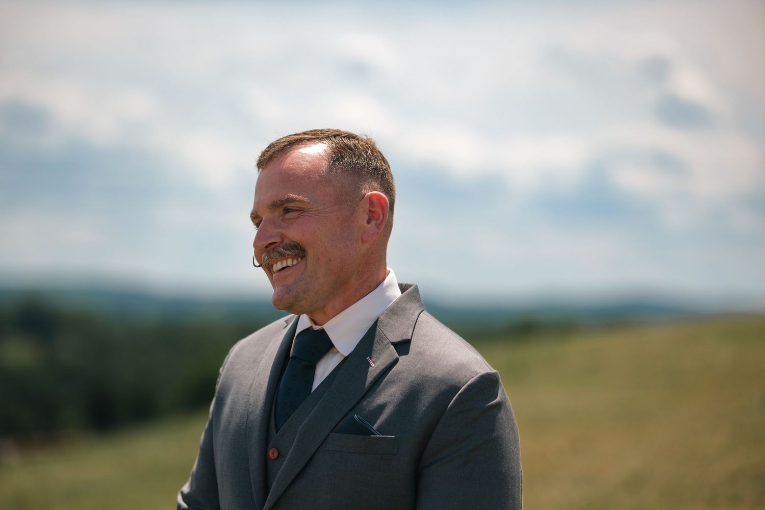 Smiling man in gray suit with mustache standing outdoors in green countryside under cloudy sky.