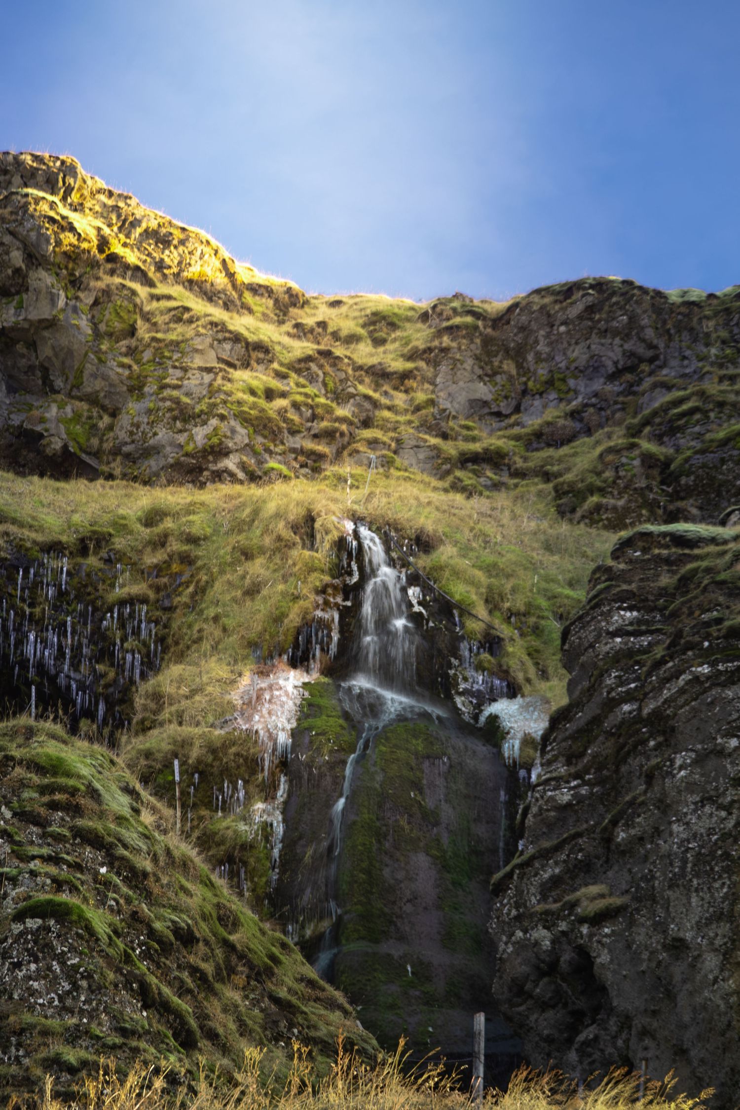 A cascading waterfall flows down a rugged cliff face surrounded by green vegetation in a mountainous landscape.