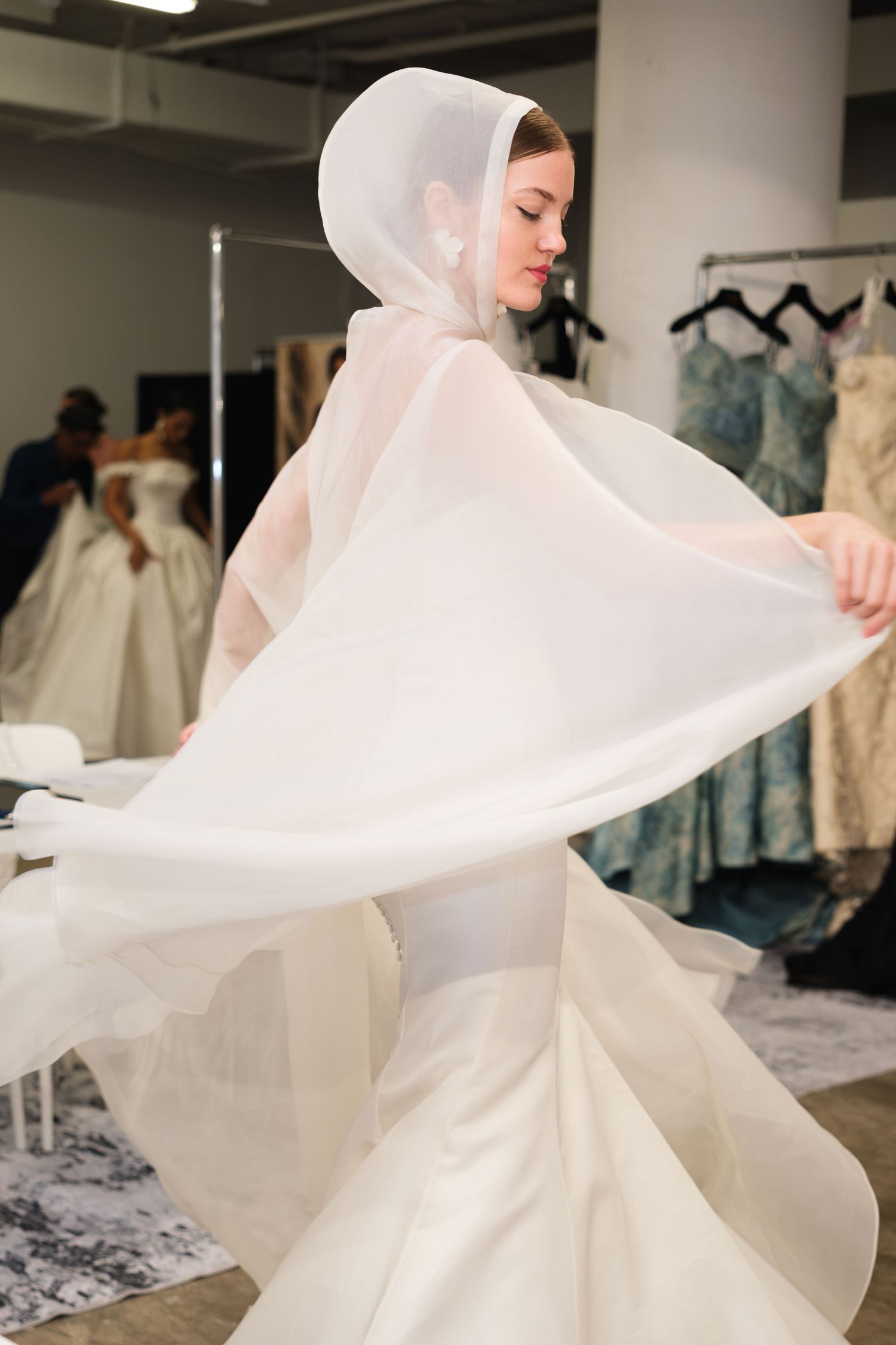 A model twirls in a white bridal gown with a flowing hooded cape in a wedding dress boutique.