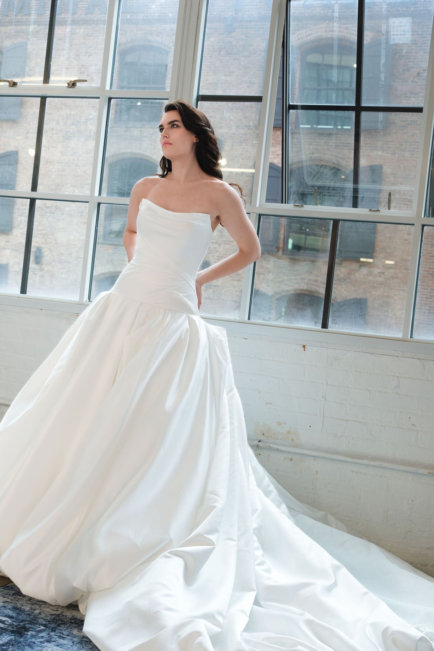 Bride in strapless white ball gown with long train poses by large industrial windows in a bright loft space.