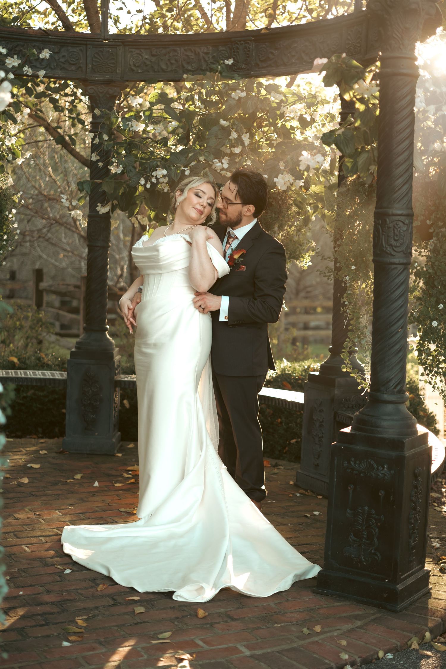 Bride in a flowing ivory satin gown embraces her groom in a dark suit beneath an ornate iron gazebo on a brick patio at an outdoor wedding.