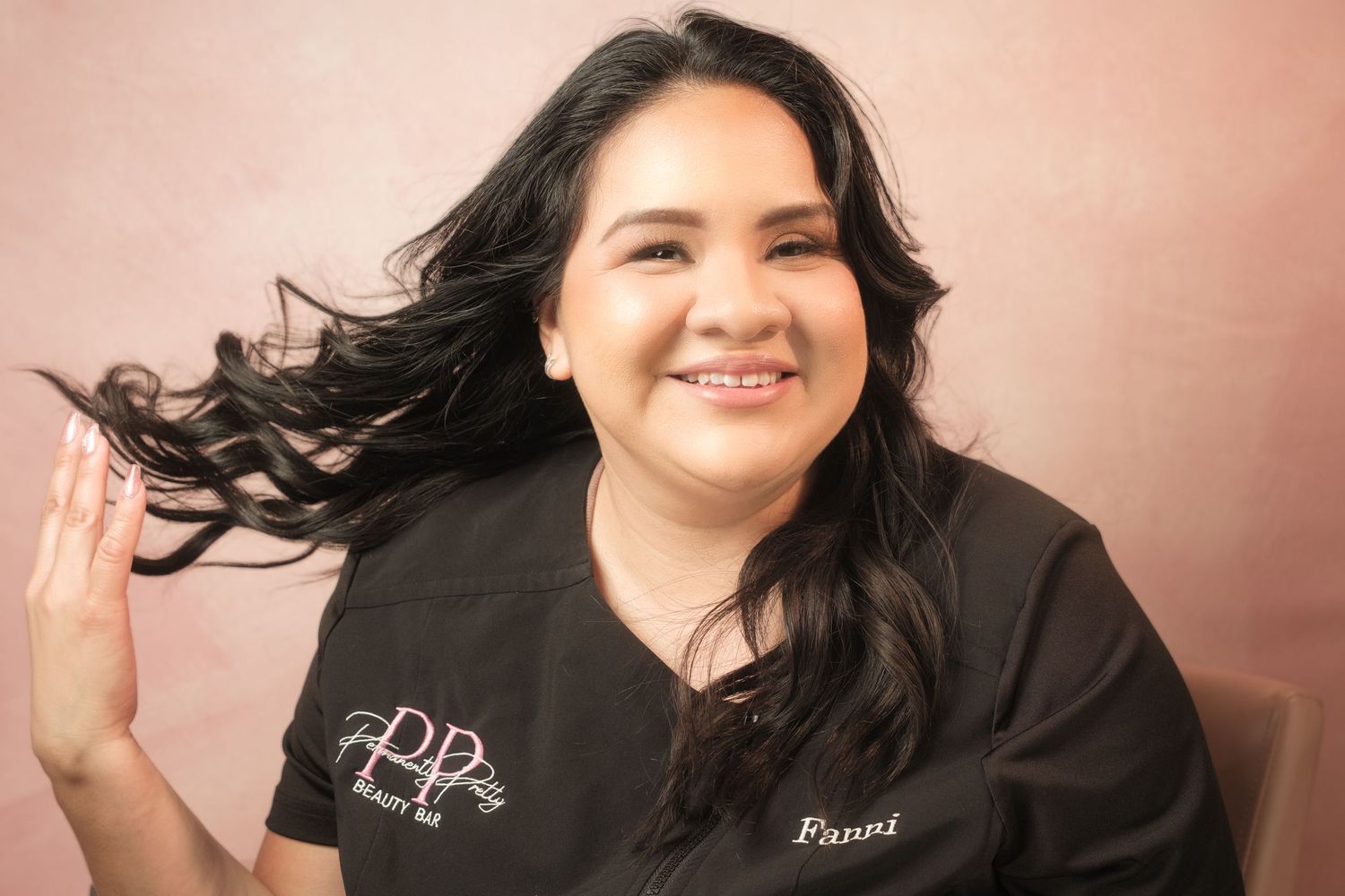 Woman named Fanni in black scrubs smiling and tossing her long dark hair in a beauty salon setting.