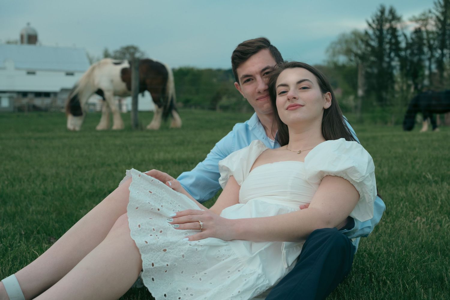 Couple sitting together on green grass with horses grazing near a white barn in a peaceful countryside setting.