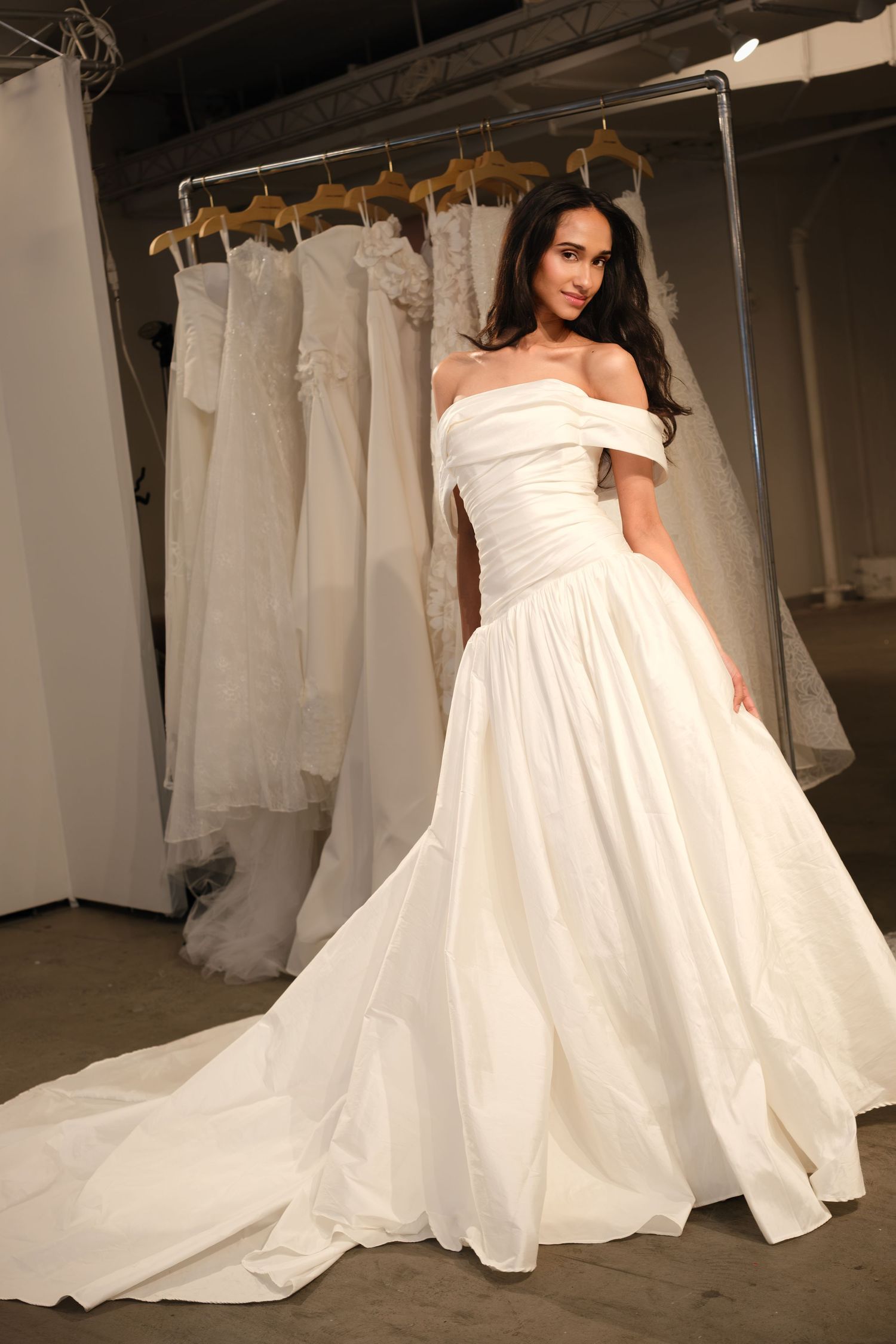 Woman in an off-the-shoulder ivory taffeta ball gown wedding dress with a long train, standing before a rack of bridal gowns.