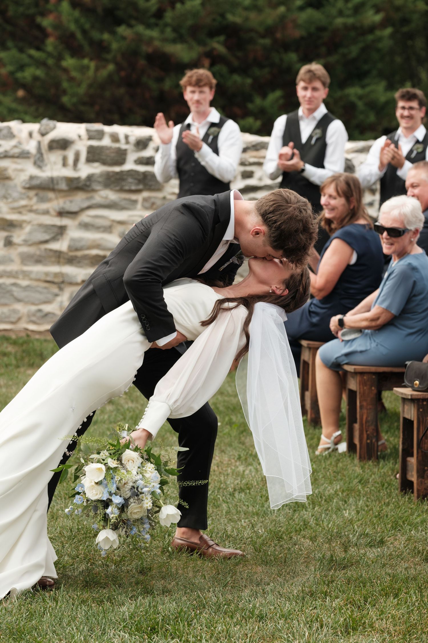 Groom dips bride for a romantic kiss at outdoor wedding ceremony while guests applaud and cheer.