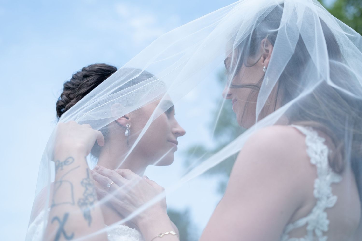 Two brides share an intimate moment under a flowing white veil on their wedding day, one with a visible tattoo.
