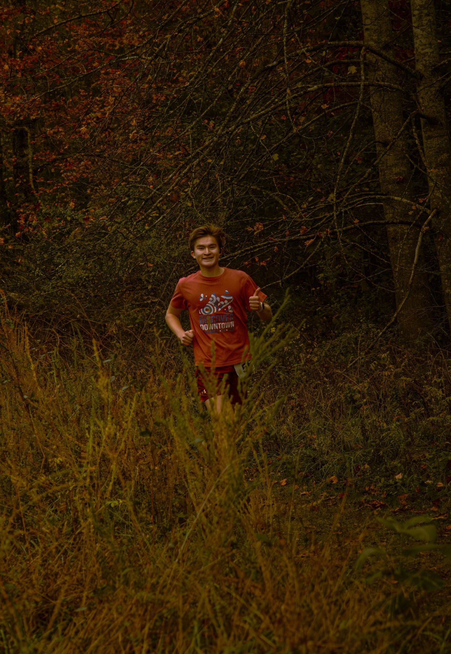 A person wearing an orange shirt stands in tall grass and brush during autumn at dusk.