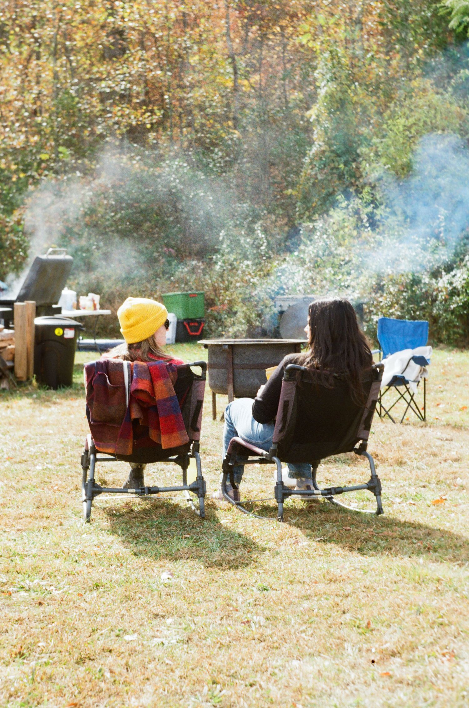 People relaxing in camping chairs under autumn trees with smoke from a campfire drifting through the scene.