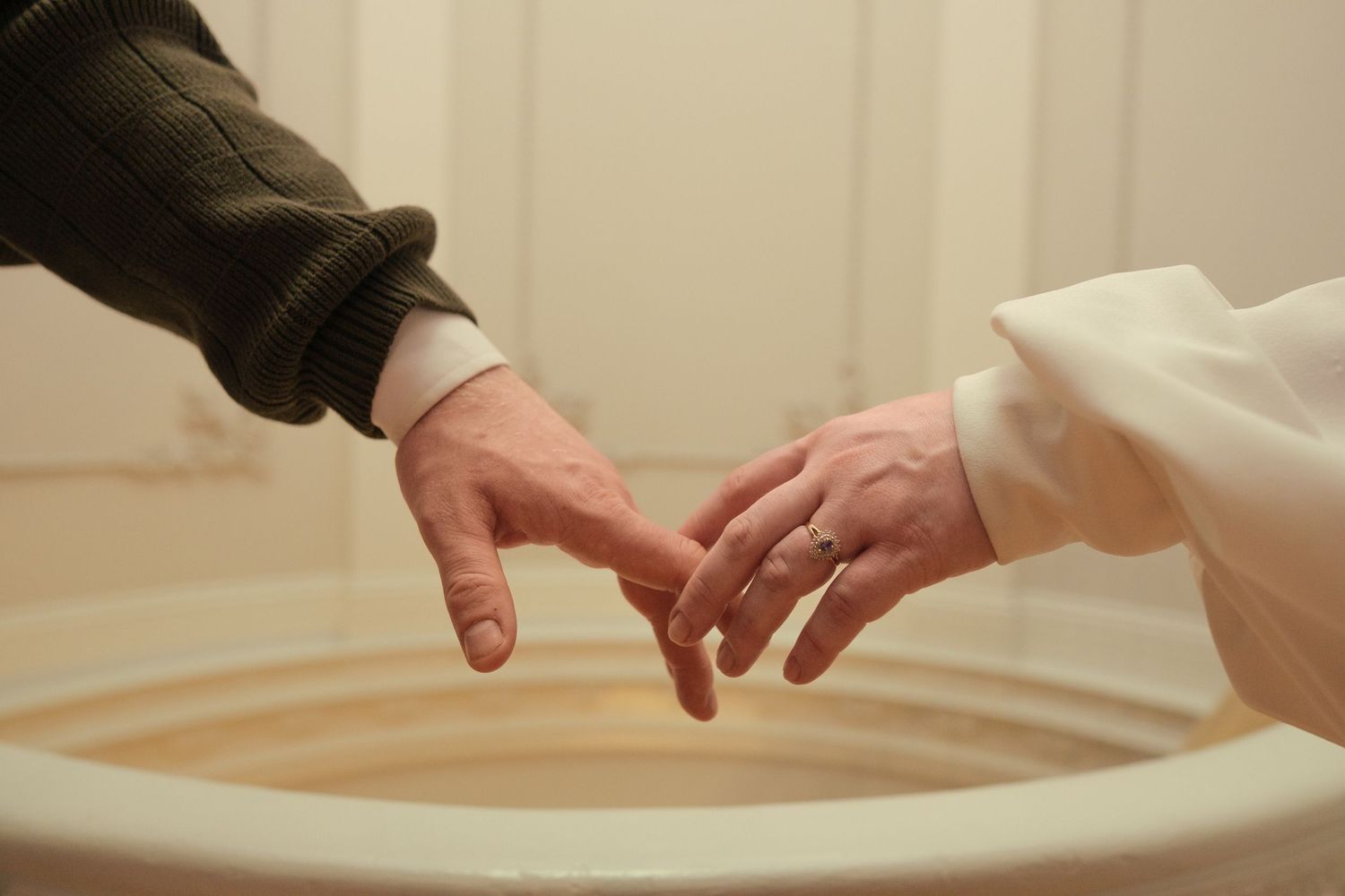 Two hands touching fingertips over a baptismal font, one in casual wear and one in white clergy robes.