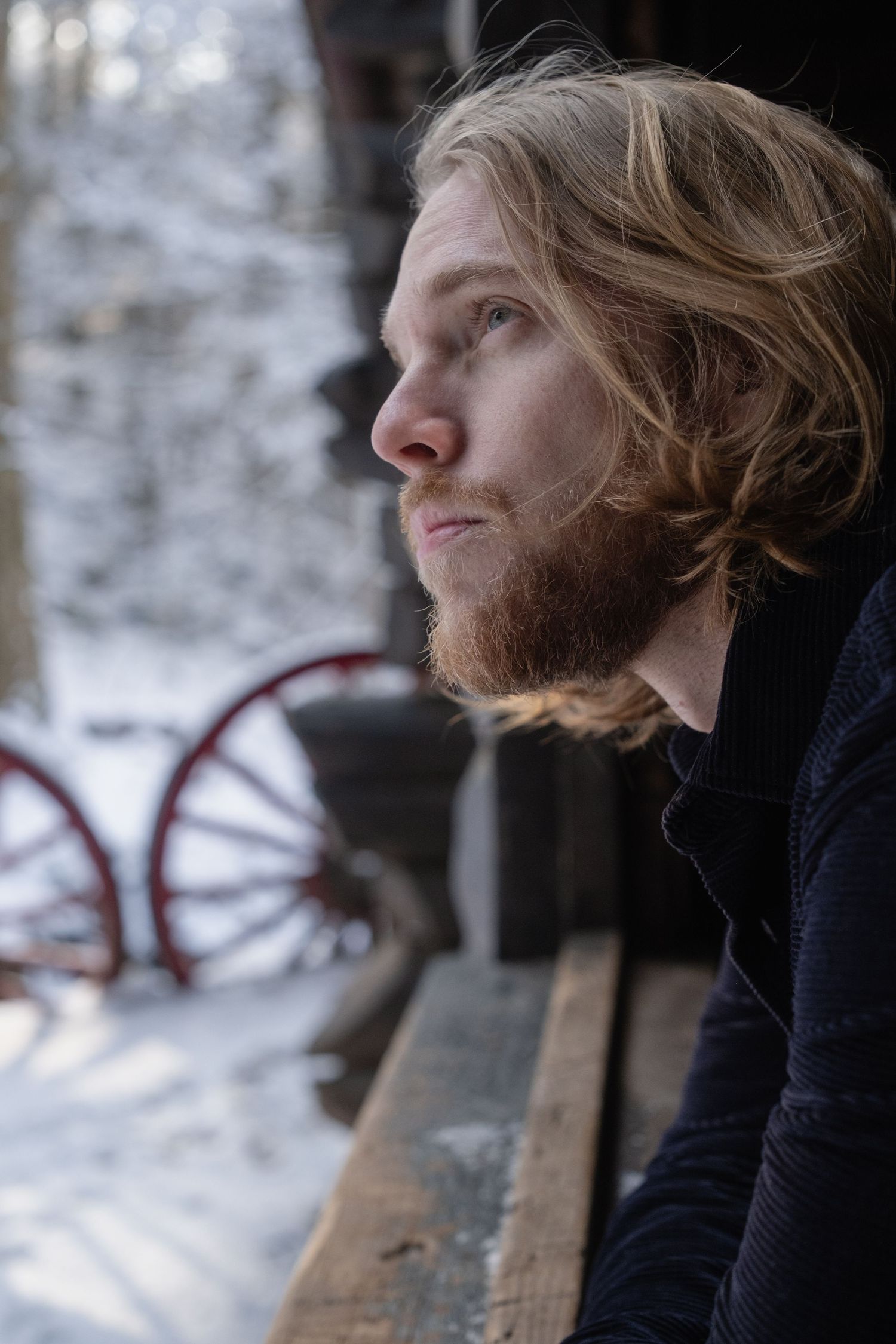 A person with long hair and beard gazes thoughtfully into the distance while sitting on a wooden bench in winter.