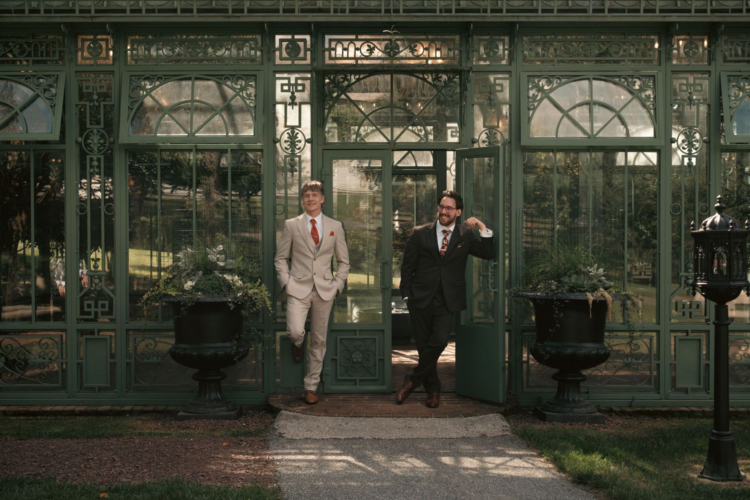 Two men in suits exit an elegant green glass conservatory building surrounded by lush gardens.