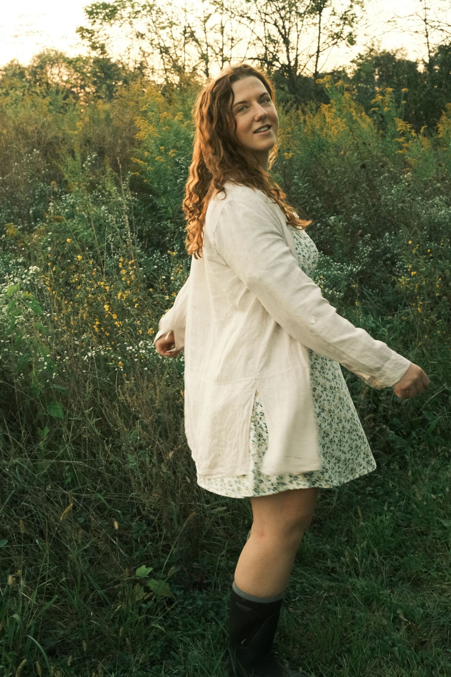 A smiling woman in a floral dress, white cardigan, and rain boots stands in a lush green field at golden hour.