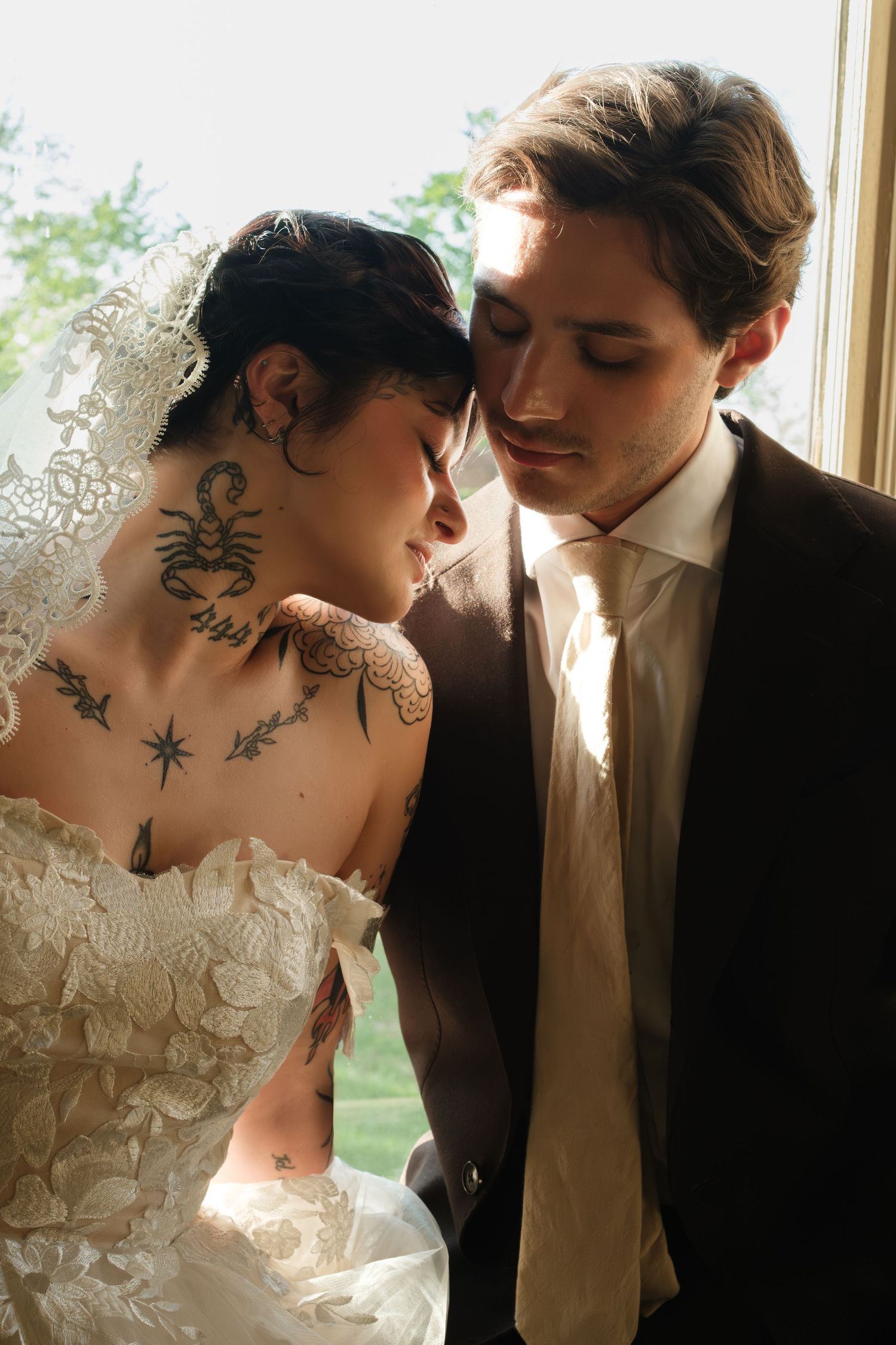 Tattooed bride in lace wedding dress and veil leans close to groom in dark suit near window.