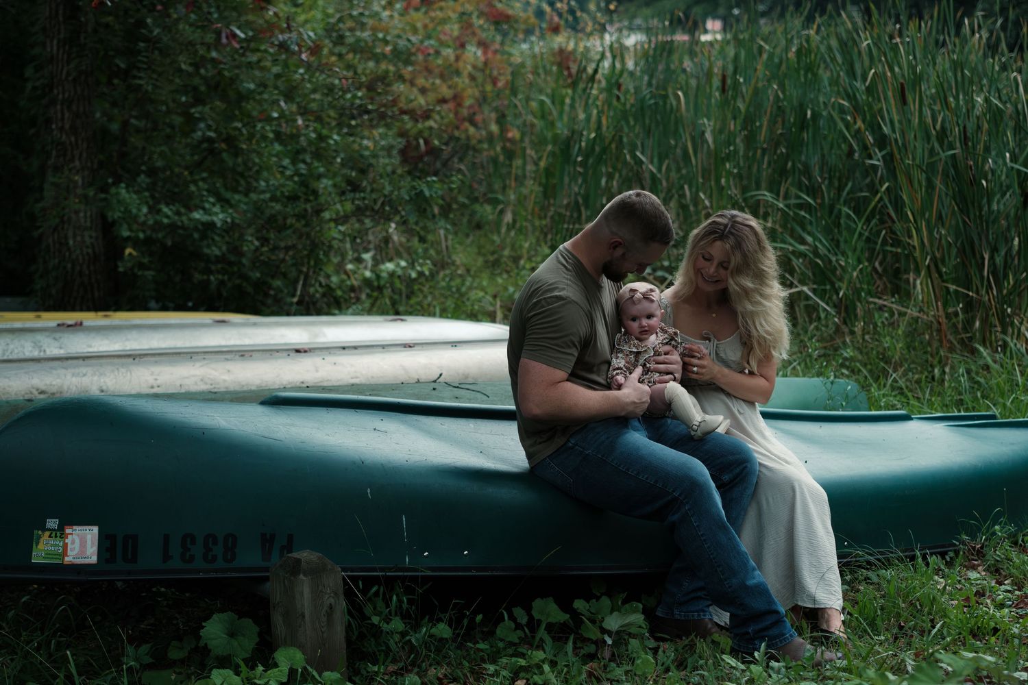 A young family sits together on a green canoe near a lush forest, parents lovingly holding their baby outdoors.