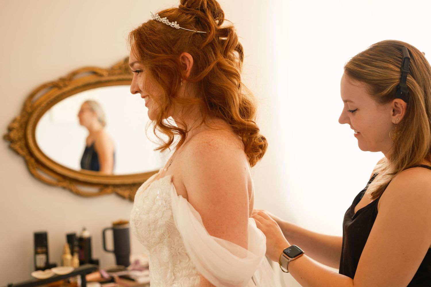 Bridesmaid helps red-haired bride button her wedding dress while reflected in ornate gold mirror.