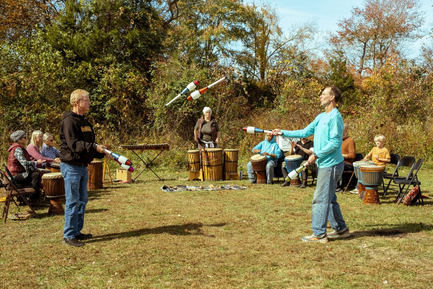 People juggling clubs outdoors in a park during autumn with spectators watching from chairs.