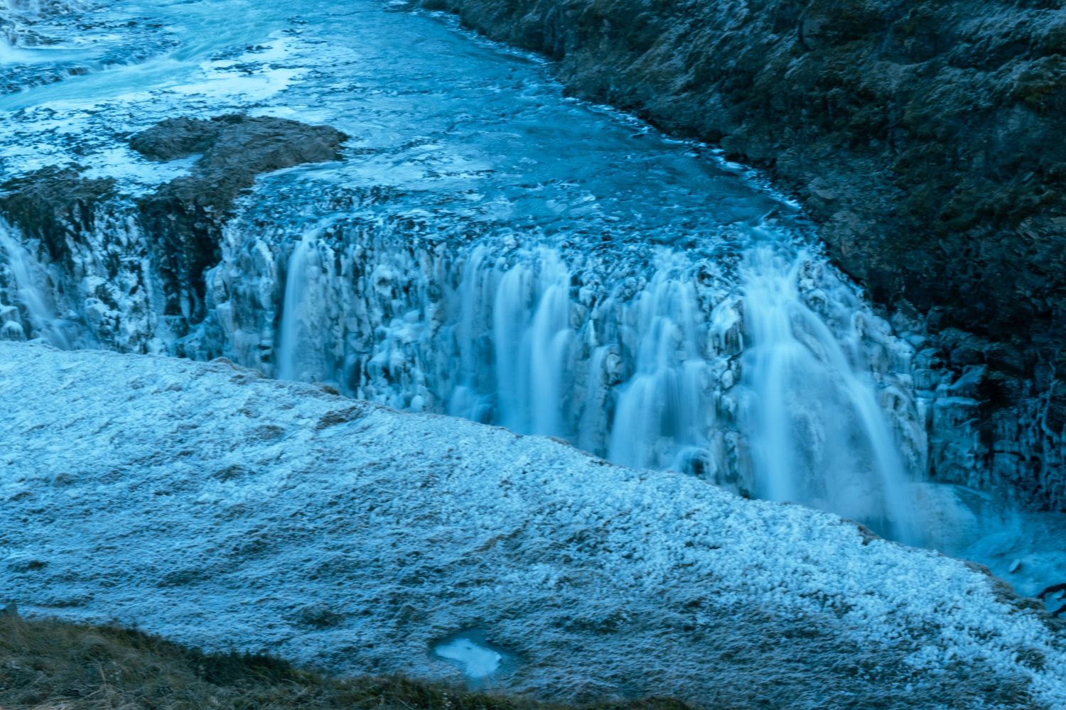A panoramic view of frozen waterfalls cascading down a rocky cliff face in winter with blue-tinted ice formations.