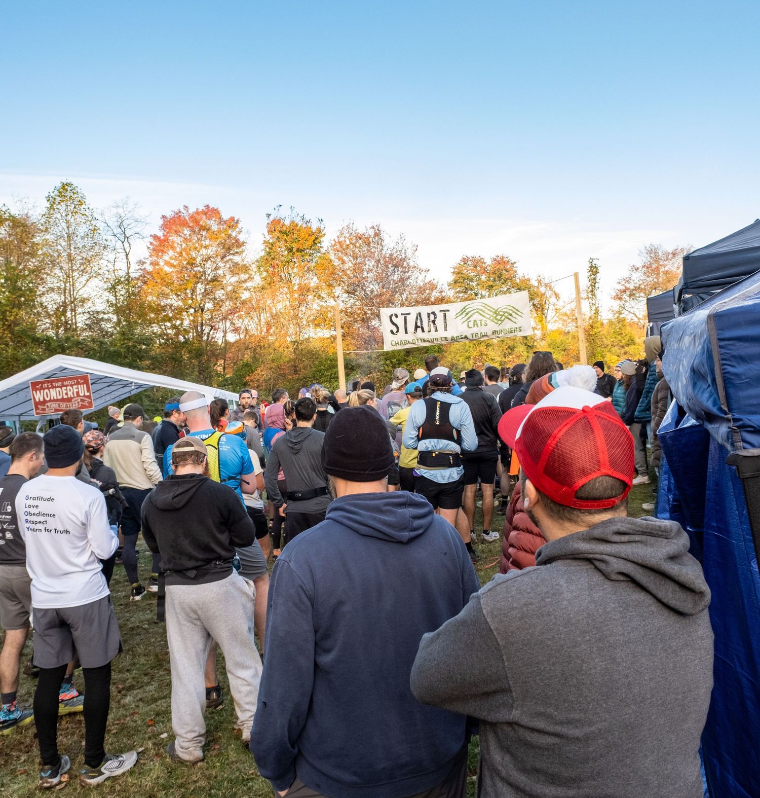 Crowd of runners gathering at a race start line on a crisp autumn morning with colorful fall foliage in the background.
