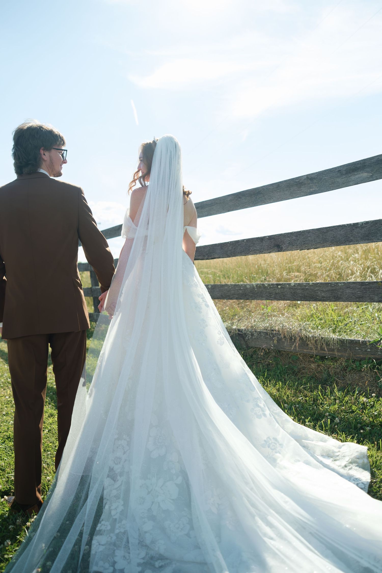 Bride in lace gown with long veil and groom in brown suit by rustic fence.