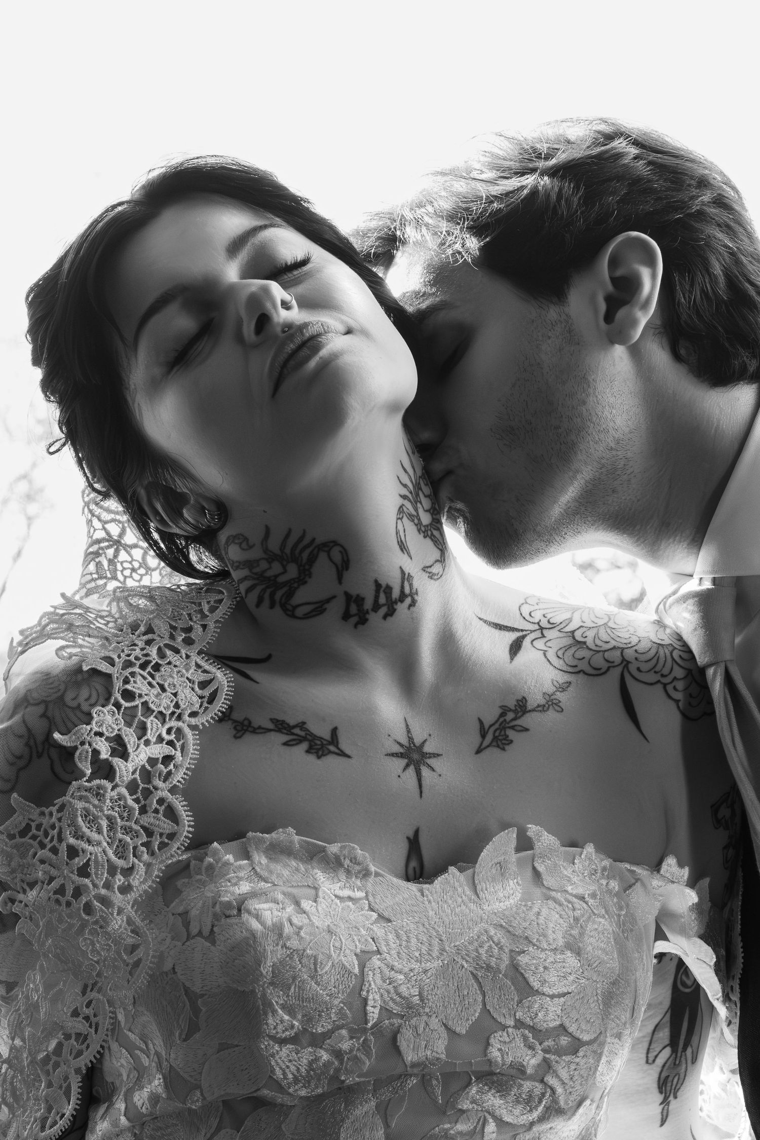 Black and white photo of a tattooed bride with closed eyes as groom kisses her neck in a romantic wedding moment.